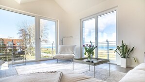 Moderni olohuone, jossa on sohva, nojatuoli, sohvapöytä ja suuret ikkunat, joista avautuu näkymä merelle.