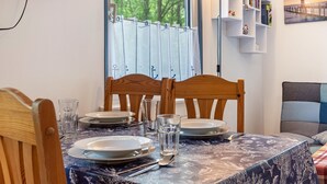 A dining area with a table set for two, chairs, a television mounted on the wall, a window with curtains, and a bookshelf with books and decorative items.