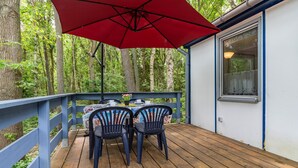A wooden deck with a table and chairs, a red umbrella, and a white building with a window.