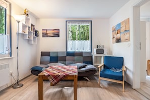 Una sala de estar acogedora con un sofá, un sillón azul, una mesa con mantel de cuadros, una estantería, una ventana con cortinas, un cuadro y un televisor pequeño.