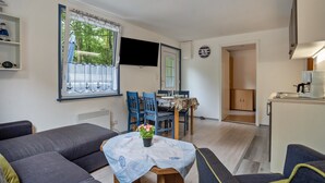 A living room with a sofa, a dining table with chairs, a TV mounted on the wall, and a kitchen area with cabinets and a refrigerator. - Bungalow 'Bungalow Kay, Bgl  4' with Private Terrace and Wi-Fi (Dranske)