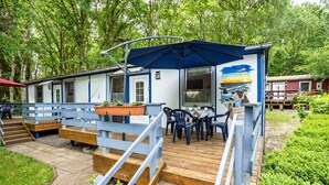A wooden deck with outdoor seating, a covered area with an umbrella, and a building with a mural. - Bungalow 'Bungalow Kay, Bgl  4' with Private Terrace and Wi-Fi (Dranske)