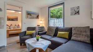 A modern living room with a sofa, armchairs, a small table with a plant, a bookshelf, and a window with a view of trees. - Bungalow 'Bungalow Kay, Bgl  4' with Private Terrace and Wi-Fi (Dranske)