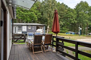 Une terrasse en bois avec une table et des chaises, un petit bâtiment orné d’un symbole d’ancre et un parasol rouge.