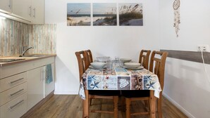 Un comedor con una mesa dispuesta para cuatro, sillas de madera, una pared blanca con tres cuadros con motivos playeros y una decoración de atrapasueños.