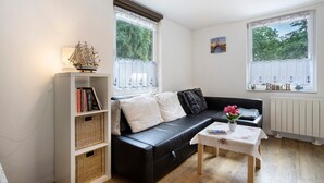 Una sala de estar con un sofá de cuero negro, una estantería blanca, una mesita con un libro y una flor, y dos ventanas con cortinas.