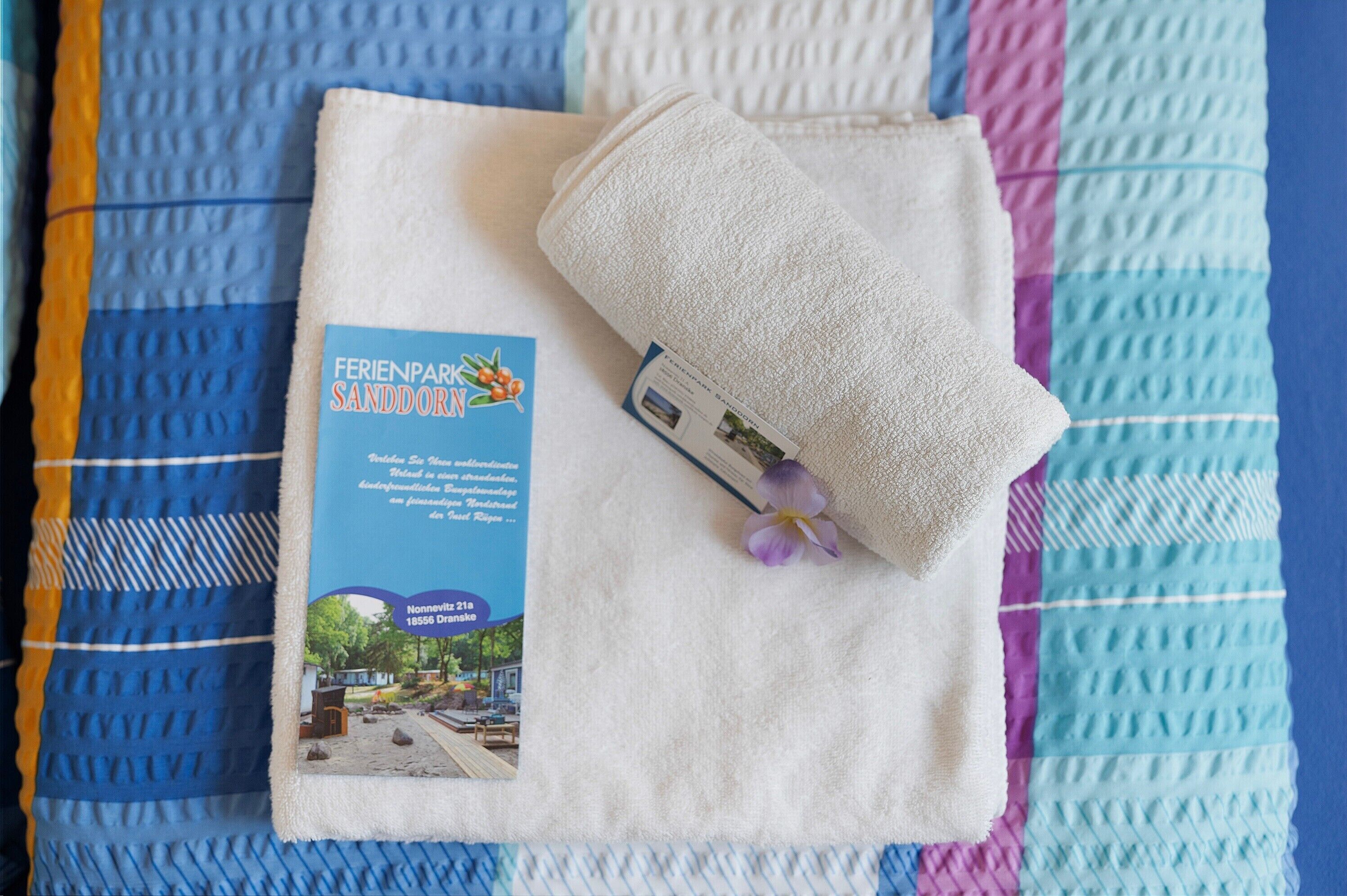 Een gevouwen witte handdoek op een bed, met een brochure getiteld 'FERIENPARK SANDDORF' en een paarse bloem.