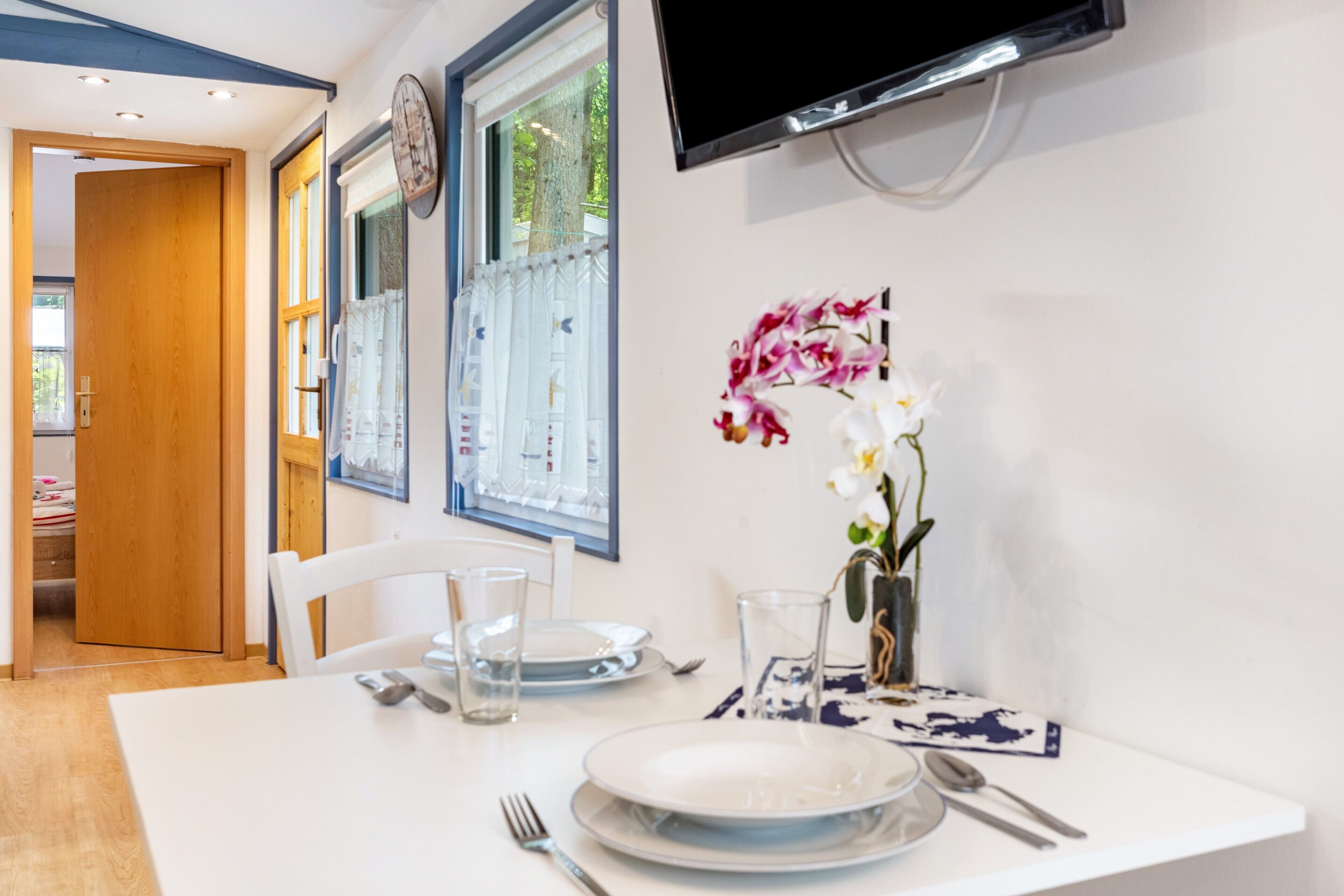 Et spiseområde med et hvidt bord, tallerkener, bestik, en vase med blomster, et ur og et TV ophængt på væggen.