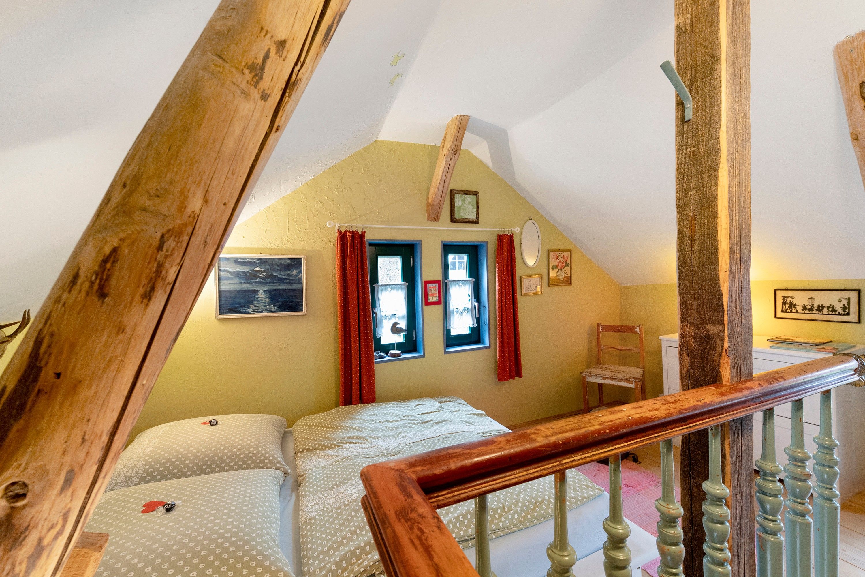 Une chambre avec un plafond en pente, un lit avec une literie à motifs, une fenêtre avec des rideaux et une rampe d’escalier en bois.