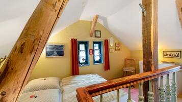 Une chambre avec un plafond en pente, un lit avec une literie à motifs, une fenêtre avec des rideaux et une rampe d’escalier en bois.