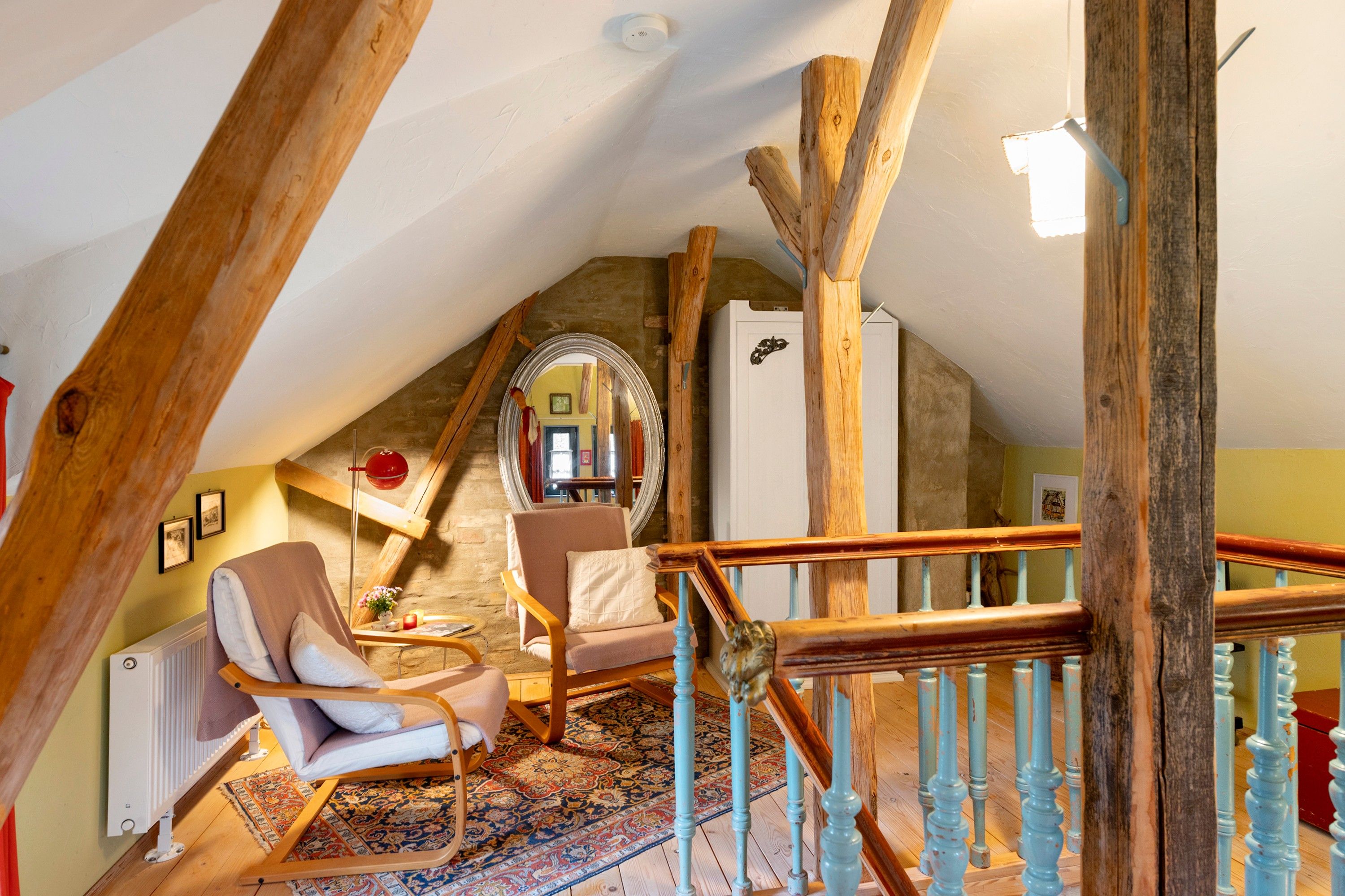 A cozy attic room with a sloped ceiling, wooden beams, a chair, a small table, a mirror, and a staircase with a wooden railing.