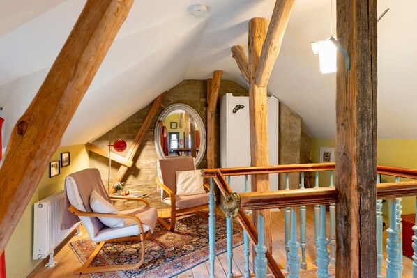 A cozy attic room with a sloped ceiling, wooden beams, a chair, a small table, a mirror, and a staircase with a wooden railing.