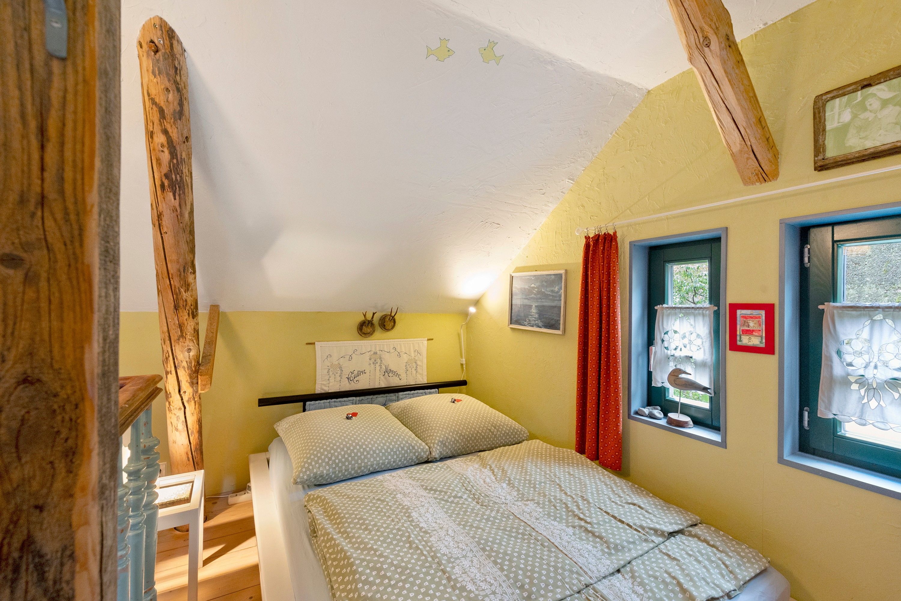 Een slaapkamer met een bed, een raam met gordijnen, een aan de muur bevestigd schilderij en een houten balkenconstructie.