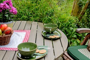 A wooden table with a green bowl, a teapot, and a cup, accompanied by a basket of apples and a checkered napkin.