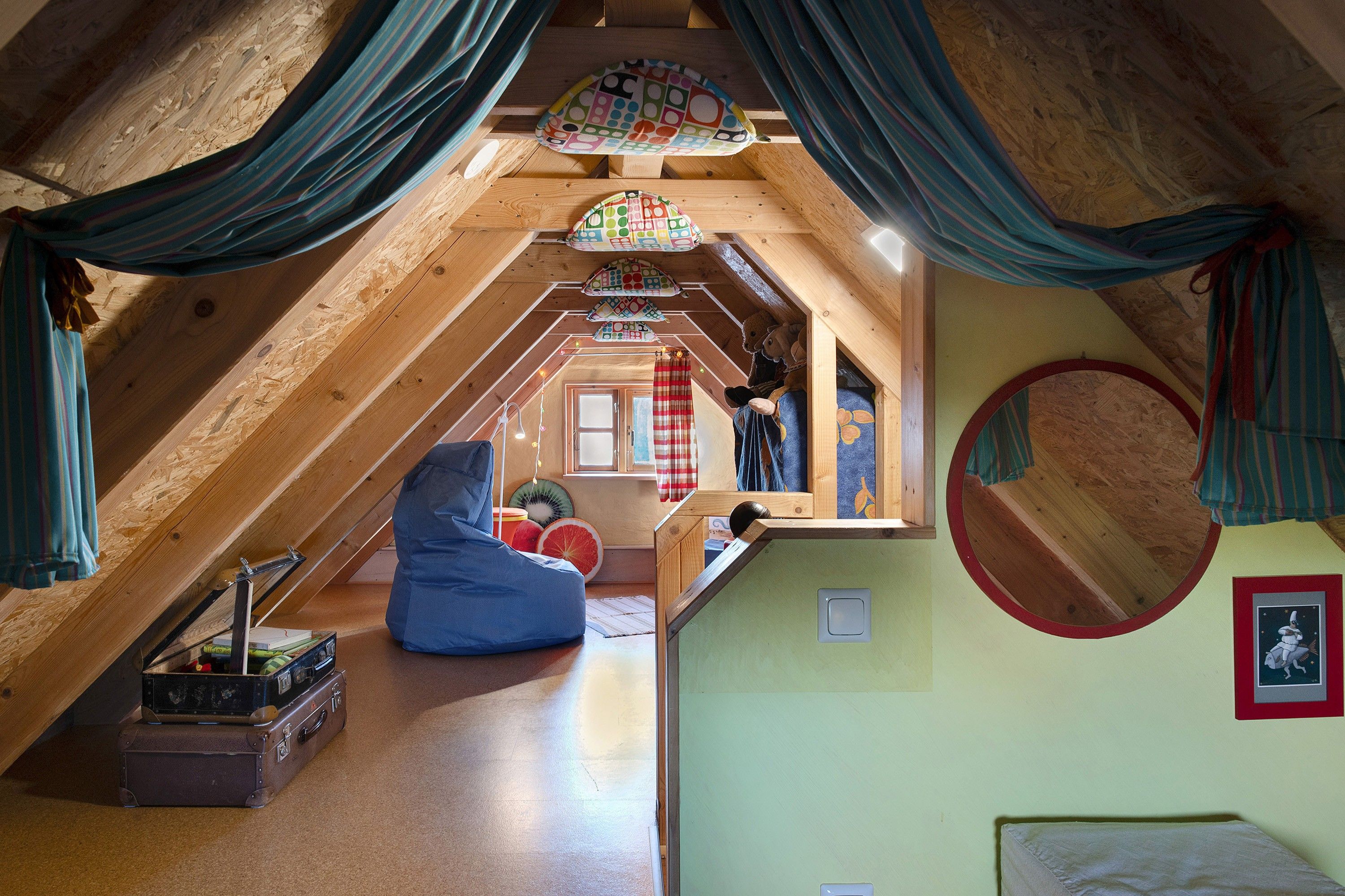 An attic bedroom with a sloped ceiling, a bean bag chair, a suitcase, and a small window with curtains.