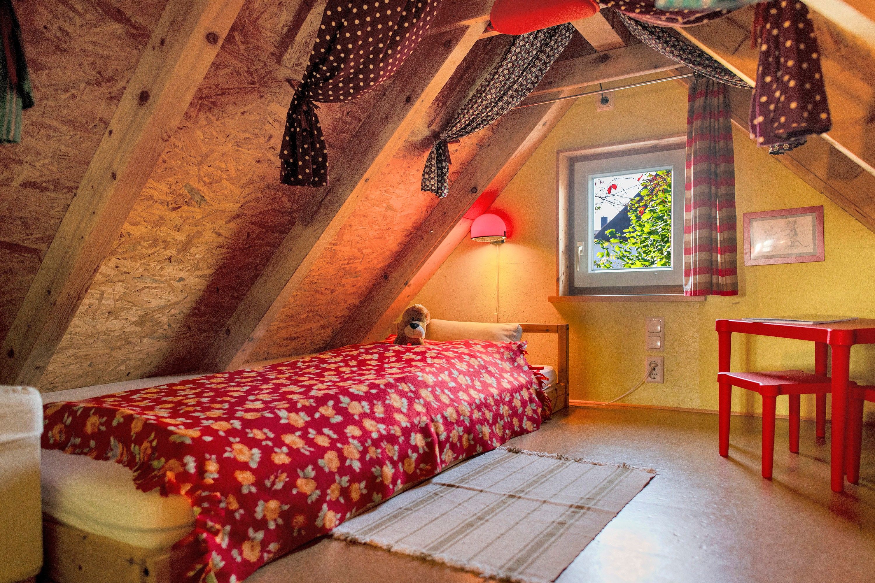 A cozy attic bedroom with a bed, a desk, and a window with a view of greenery.