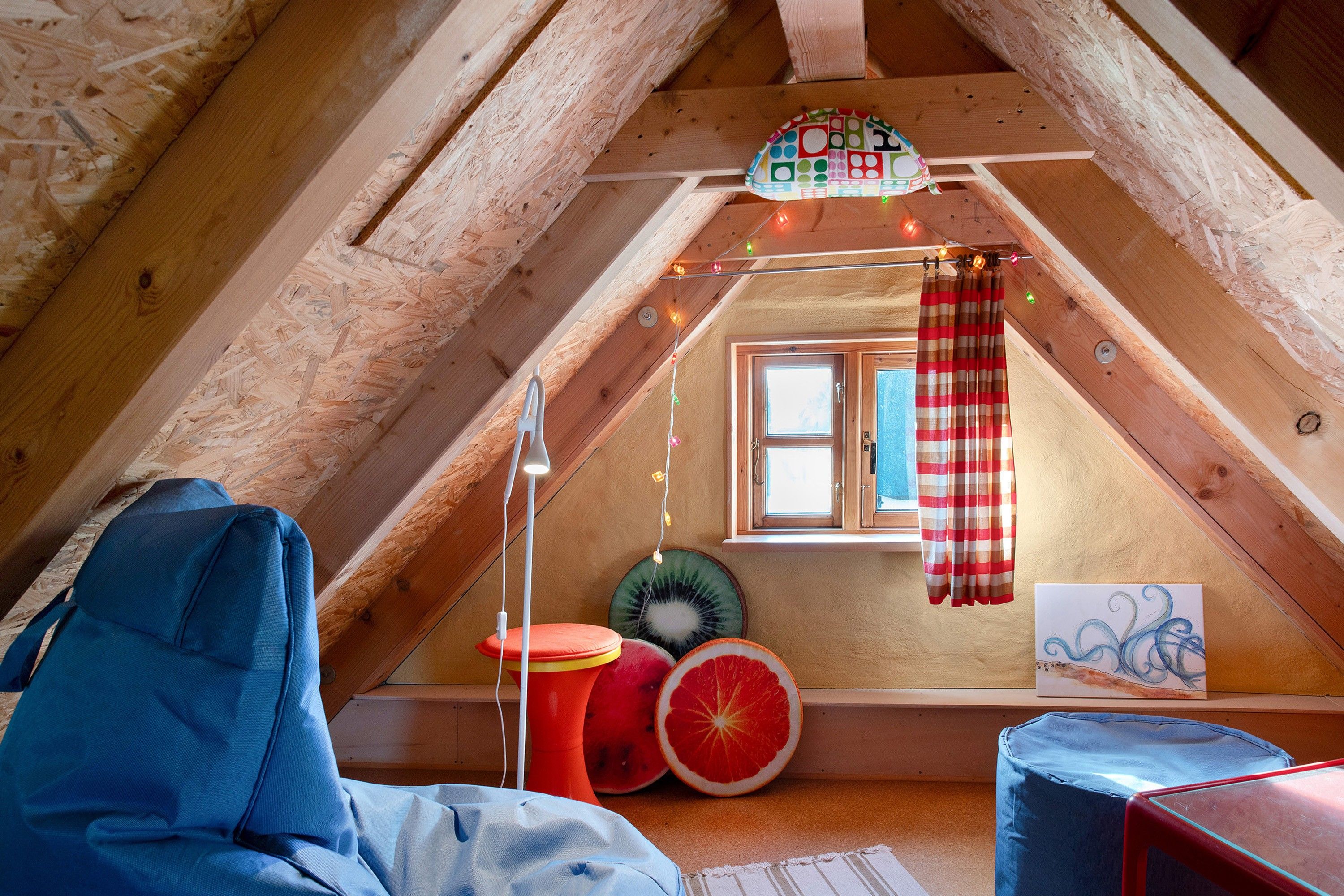 Una habitación cálida en el ático, con techo inclinado, ventana con cortinas, techo de madera y una pequeña mesa con frutero.