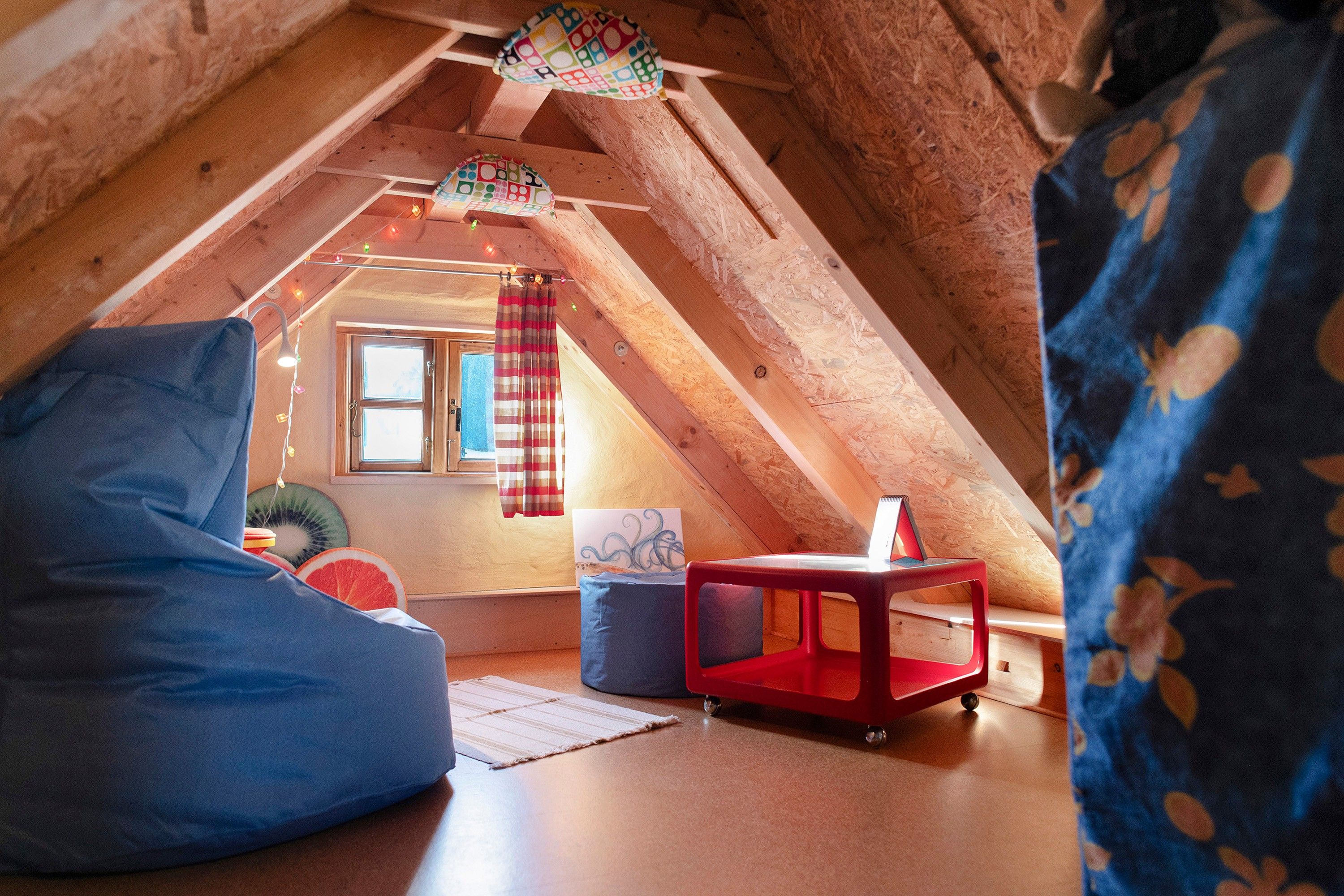 Ein gemütliches Dachzimmer mit geneigter Decke, einem Sitzsack, einem kleinen roten Tisch und einem Fenster mit Vorhängen.