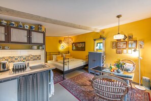 Quarto compacto com cama, uma mesinha, uma cadeira, armário e cozinha equipada com pia e utensílios.