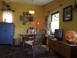 Een kamer met een blauwe kast, een rieten stoel, een bureau met een computer, een wereldbol en ingelijste foto's aan de muur.