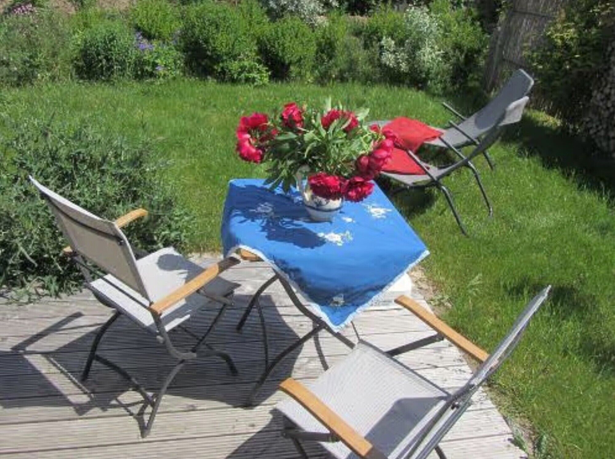 Una terraza de madera con una mesa cubierta con un mantel azul, dos sillas y un jarrón con flores rojas.