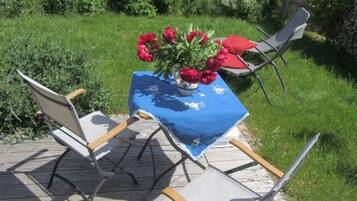 Una terraza de madera con una mesa cubierta con un mantel azul, dos sillas y un jarrón con flores rojas.
