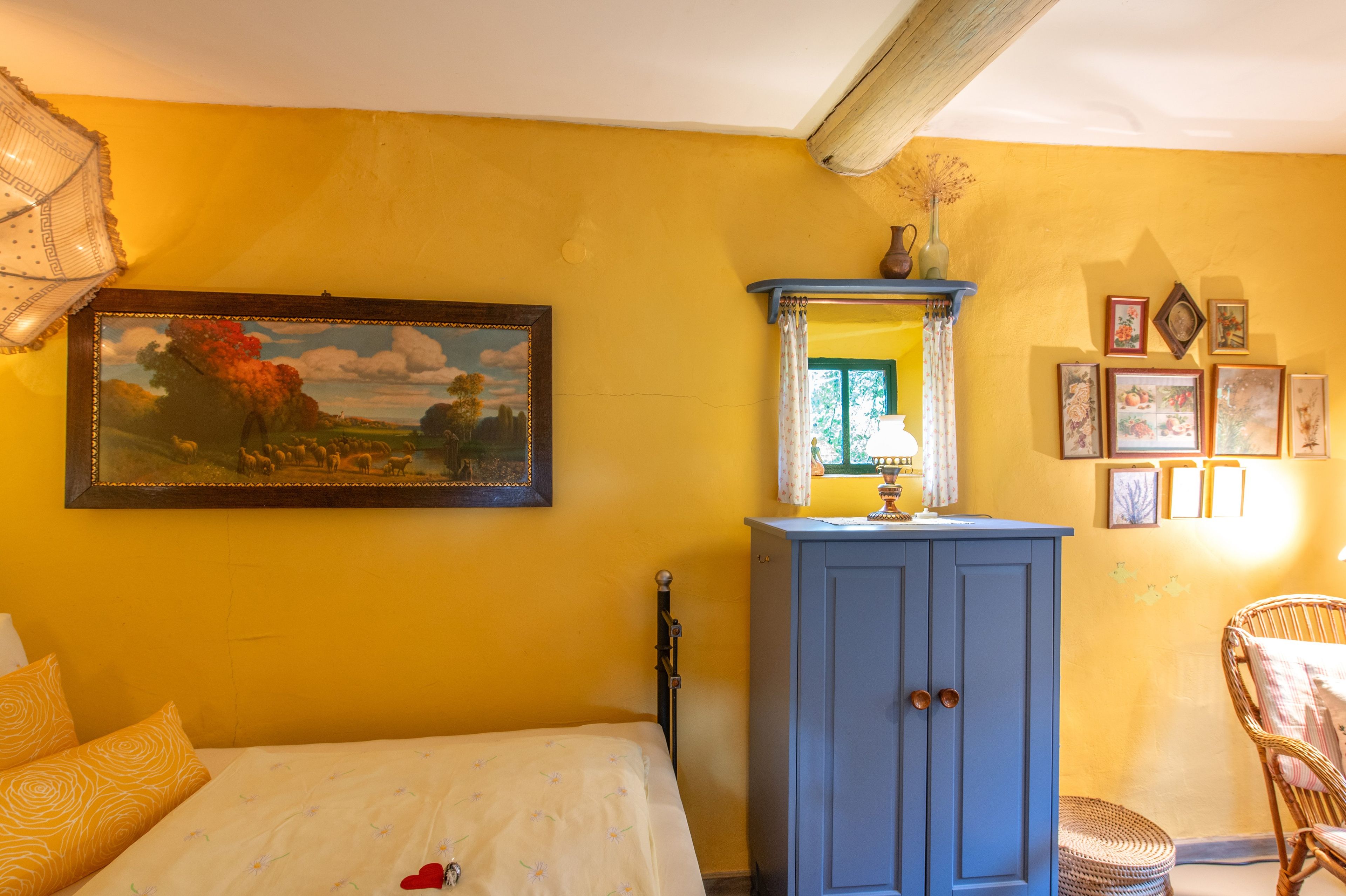 A room with a bed, a blue cabinet, a painting, and a wicker chair.