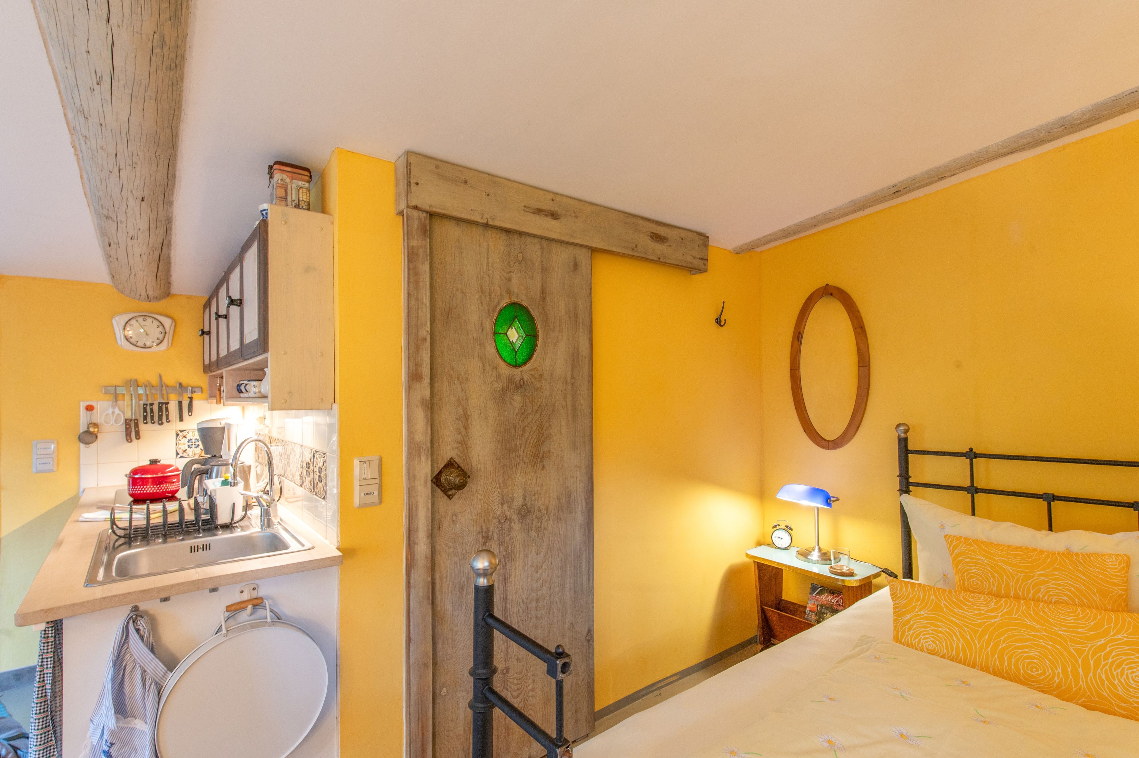 A room with a bed, a nightstand, a wall clock, a mirror, and a wooden door.