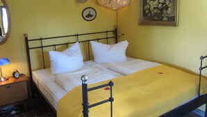 A bed with a metal frame, white bedding, a yellow blanket, a bedside table with a lamp, a wall clock, a hat, and a framed picture. - Holiday Home 'Charlottes Landlust' with Private Terrace and Garden (Ribnitz-Damgarten)