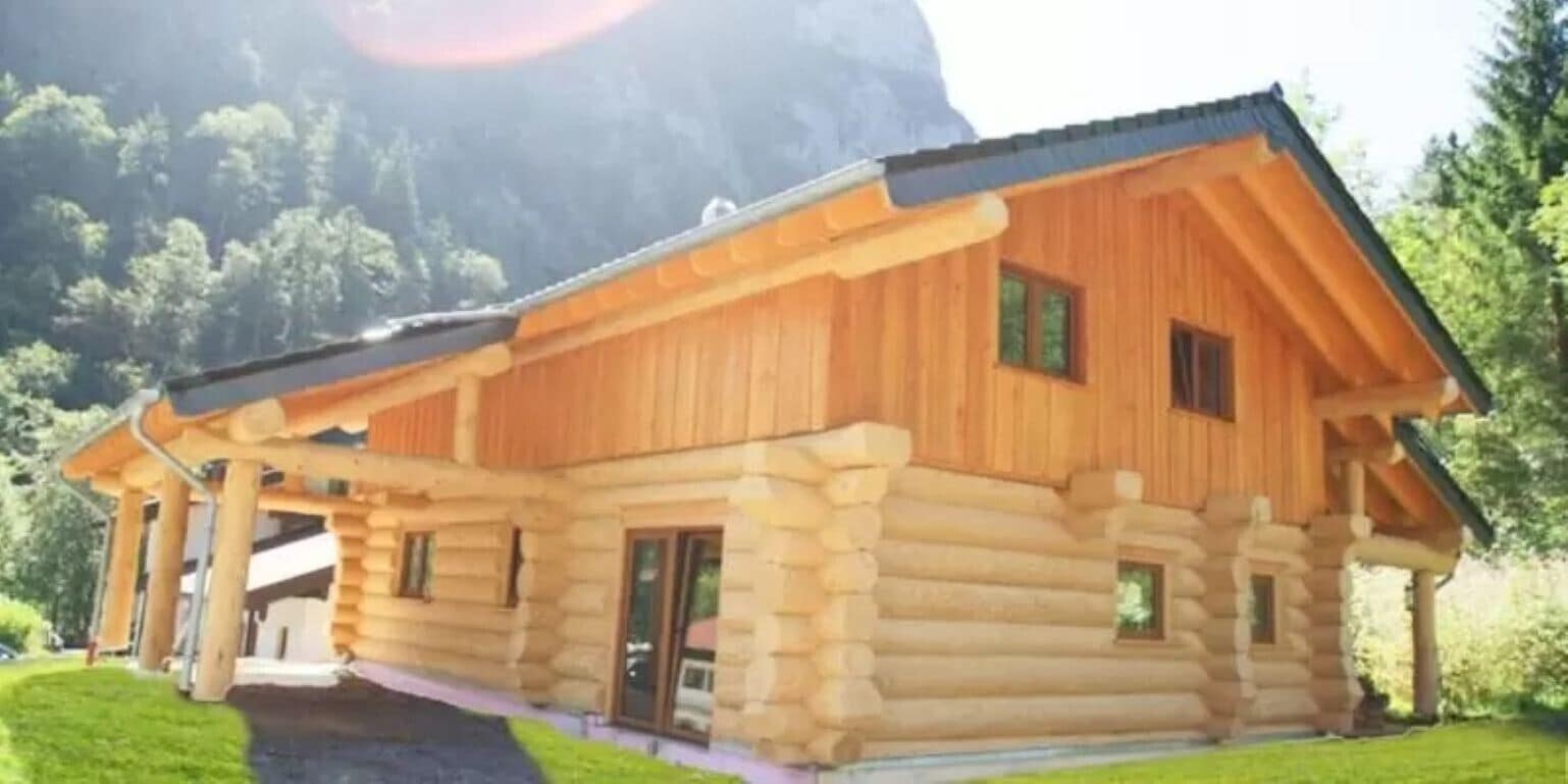 Una cabaña de troncos con un porche, una chimenea y montañas al fondo.