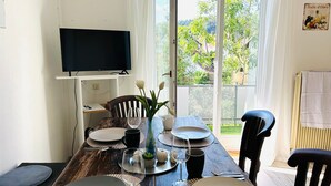 Une table à manger est dressée avec des assiettes, des verres et un vase rempli de fleurs. Un téléviseur est fixé au mur et offre une vue sur la verdure à l’extérieur.