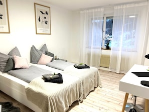Une chambre avec deux lits, une table de chevet, une fenêtre avec des rideaux légers et une œuvre d’art encadrée au mur.