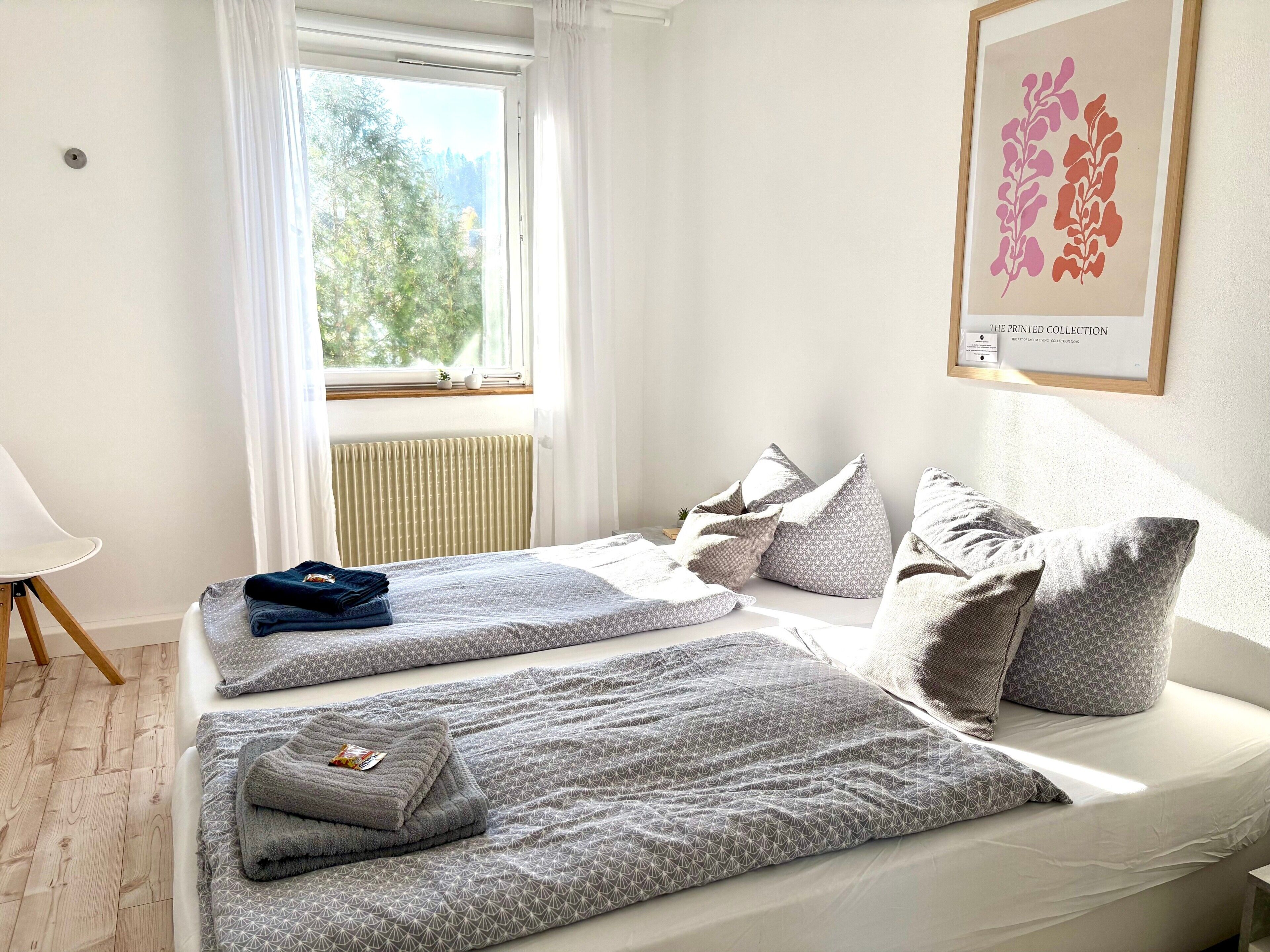 Ein Schlafzimmer mit zwei Betten, einem fensterbelaubten Fenster, einem Heizkörper und einem gerahmten Kunstwerk an der Wand.