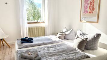 Ein Schlafzimmer mit zwei Betten, einem fensterbelaubten Fenster, einem Heizkörper und einem gerahmten Kunstwerk an der Wand.