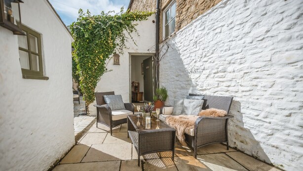 Une terrasse dotée de meubles en osier, d’une table en verre et d’un mur blanc orné d’une plante grimpante.