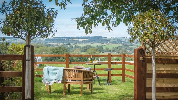 A wooden bench with a blanket, a table with a bottle and glasses, and a view of rolling hills. - Sun Hill Palins -  a cottage that sleeps 2 guests  in 1 bedroom (Leyburn)