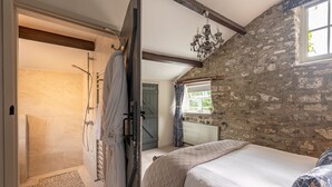 A bedroom with a stone wall, a bed with a quilted cover, a window with a view, and a shower area with a towel rack. - Sun Hill Palins -  a cottage that sleeps 2 guests  in 1 bedroom (Leyburn)