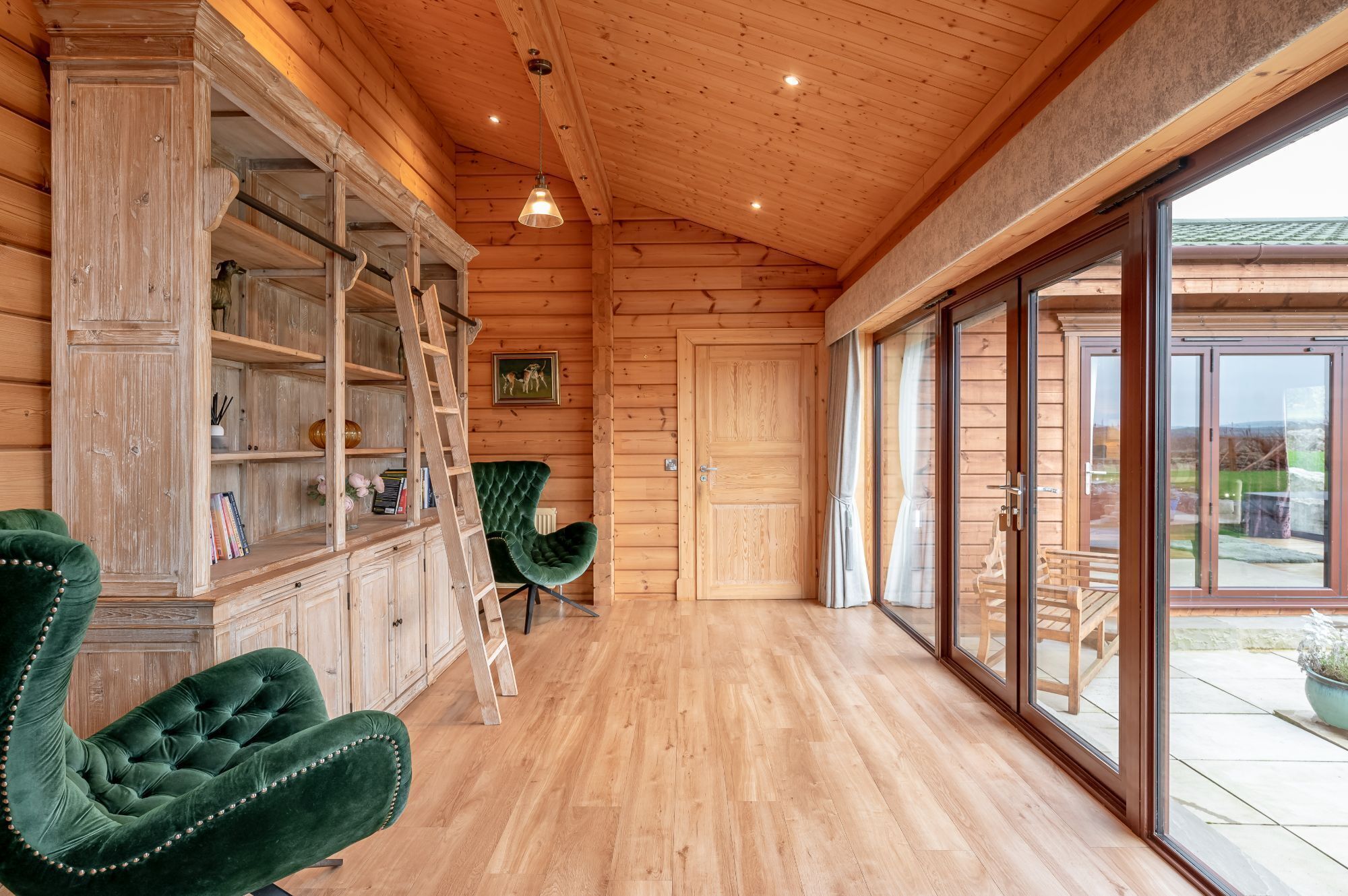 A wooden interior with a wooden floor, a green upholstered chair, a wooden cabinet, and a door leading to an outdoor area.