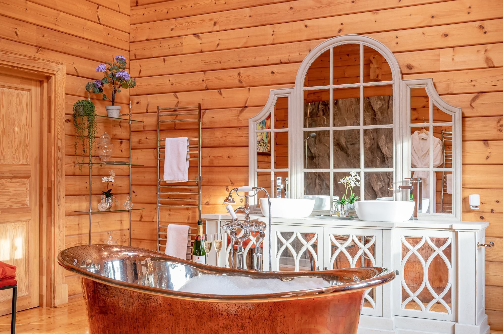 Interior de una cabaña de madera con una bañera grande, un tocador blanco con espejo y una puerta de madera.
