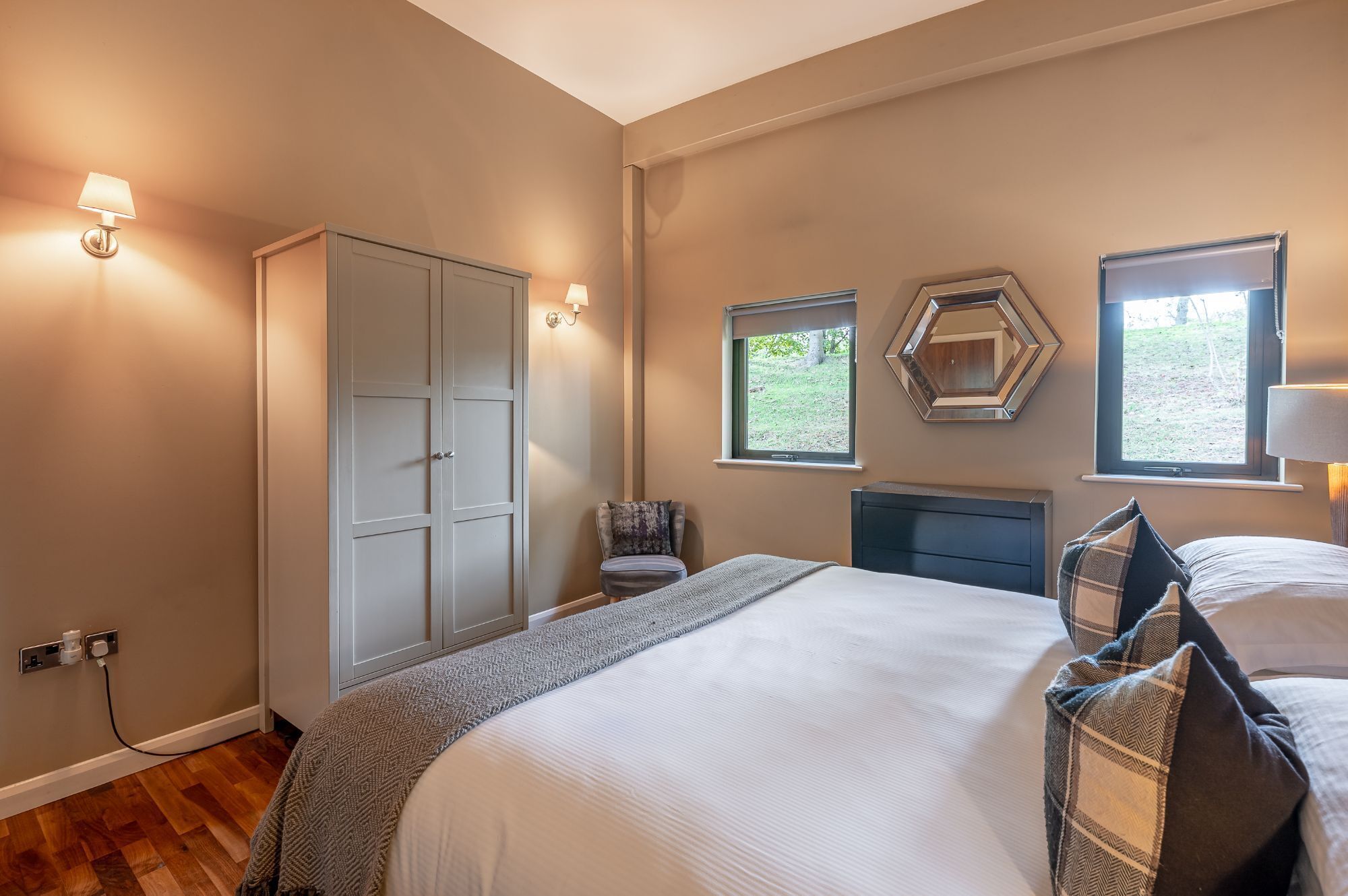 A bedroom with a bed, two bedside tables, a wardrobe, a chair, and a window with a view of greenery.