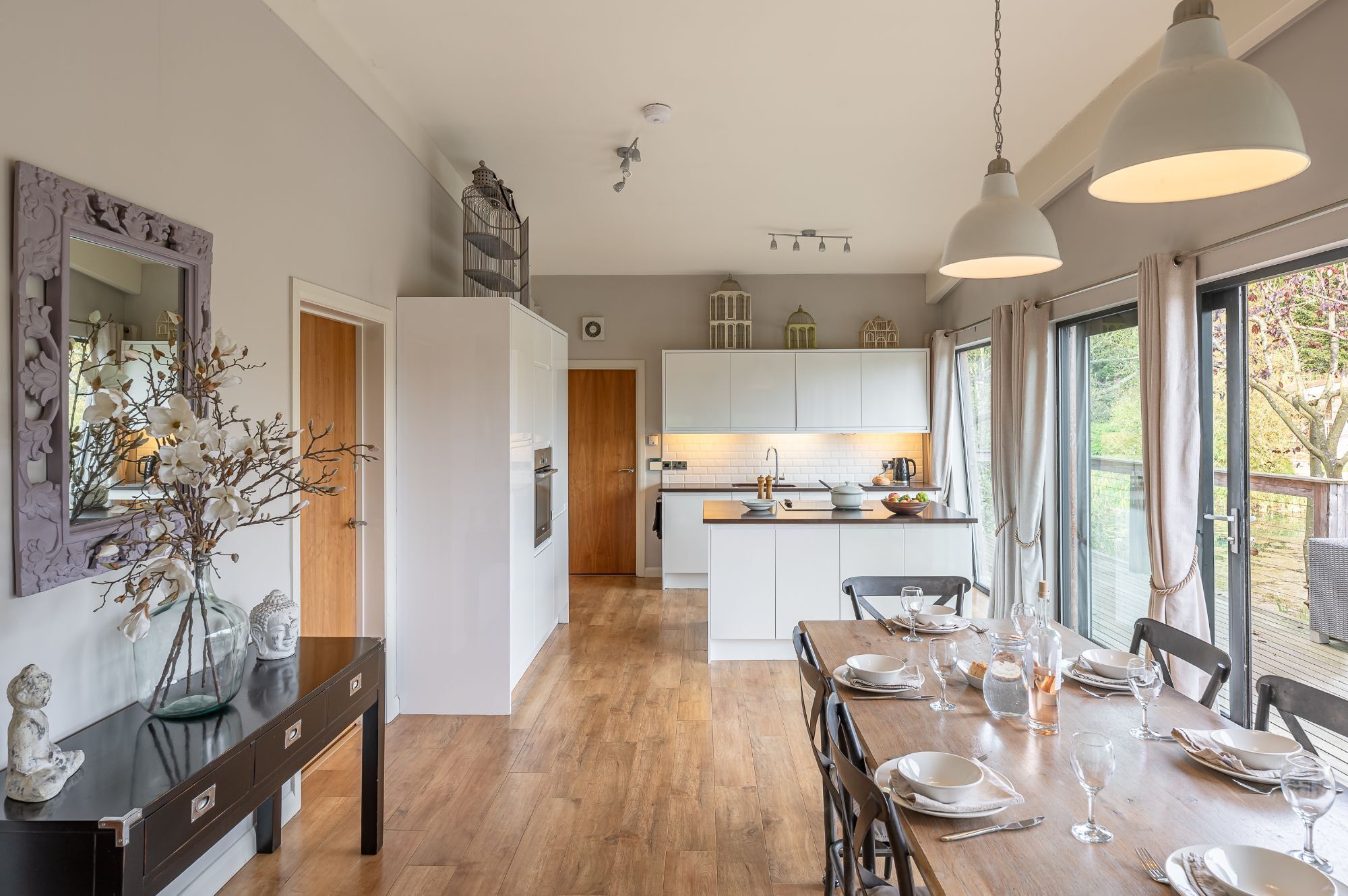 Une cuisine moderne avec des meubles blancs, une table à manger en bois pour quatre personnes et une grande fenêtre donnant sur l’extérieur.