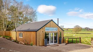 A wooden cabin with a sloped roof, large windows, and a gravel driveway. - Bearskin Cottage - Holiday At Home (Leyburn)