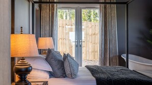 A bedroom with a bed, two bedside lamps, a bedside table with books, and a view of a wooden fence through a glass door. - Bearskin Cottage - Holiday At Home (Leyburn)