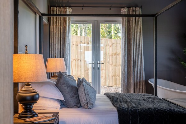 A bedroom with a bed, two bedside lamps, a bedside table with books, and a view of a wooden fence through a glass door.