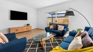 A modern living room with a blue sofa set, a glass coffee table, and a flat-screen TV mounted on the wall. - Halfway Gem - 3BR Home Between Geelong & The Coast (Mount Duneed)