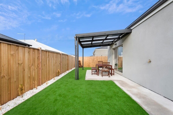 A backyard with artificial grass, a wooden fence, and a patio area with a table and chairs.
