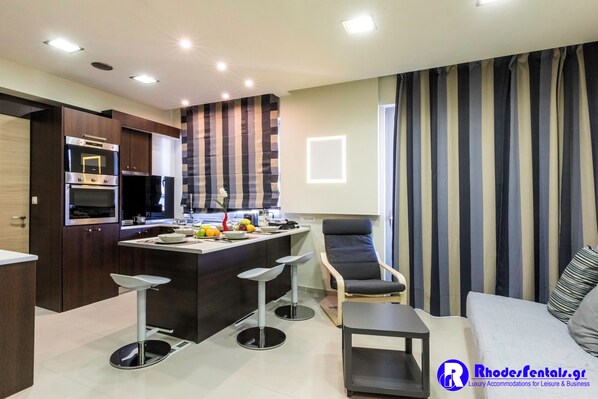 A modern kitchen with built-in appliances, a dining area with bar stools, and a living space with a sofa and striped curtains.