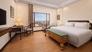Habitación de hotel con una cama grande, un escritorio con lámpara, una silla y vistas a la ciudad a través de un ventanal.