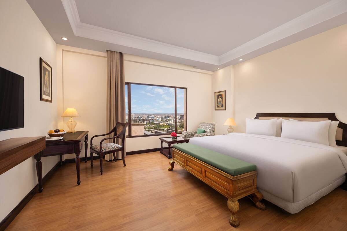 A hotel room with a large bed, a desk with a lamp, a chair, and a view of the city through a large window.