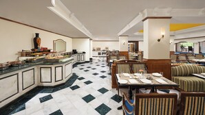 Un comedor de hotel con suelo de mármol, bufé, mesas y sillas, y vistas a la cocina.
